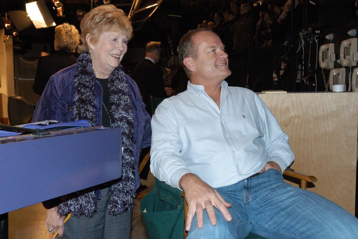 gabrielle-james-frasier Kelsey Grammer is seated in a director's chair, with Gabrielle James standing close behind him, both are smiling and looking to their left-hand side