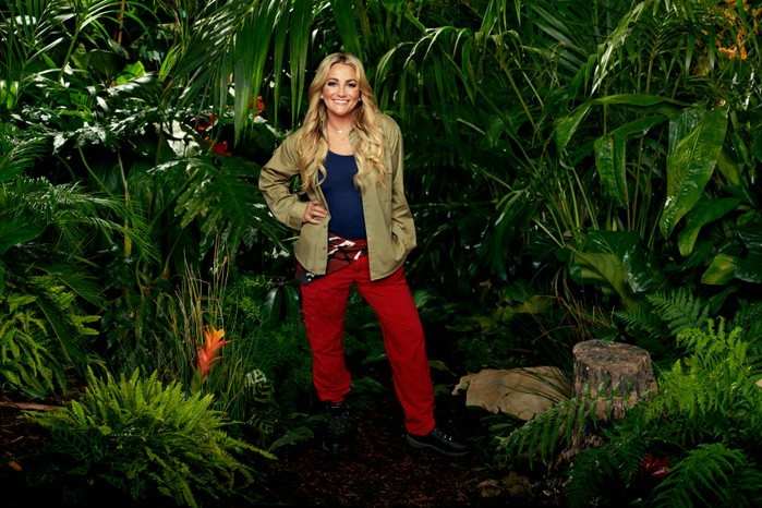 ©ITV Plc Jamie Lynn Spears wearing her I'm a Celebrity camp clothes (khaki shirt, blue vest and red trousers) while posing in front of some bushes ahead of I'm a Celebrity 2023