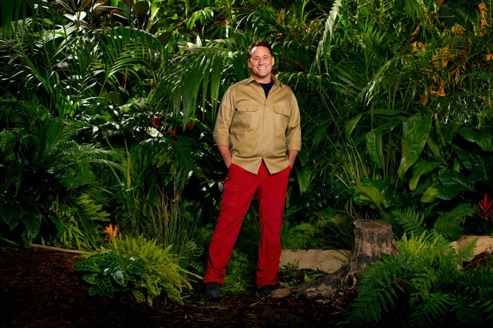 Nick-Pickard-for-IAC-2023 Nick Pickard dressed in a khaki shirt and red trousers while posing in front of some trees for I'm a Celebrity 2023.