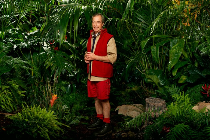 Nigel-Farage-for-I'm-a-Celebrity Nigel Farage wearing his I'm a Celebrity camp clothing (khaki shirt, red gilet, red trousers and black boots) as he poses in front of some bushes ahead of I'm a Celebrity 2023.