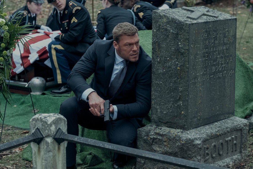 Alan Ritchson as Jack Reacher in Reacher season 2 hiding behind a tombstone, holding a pistol
