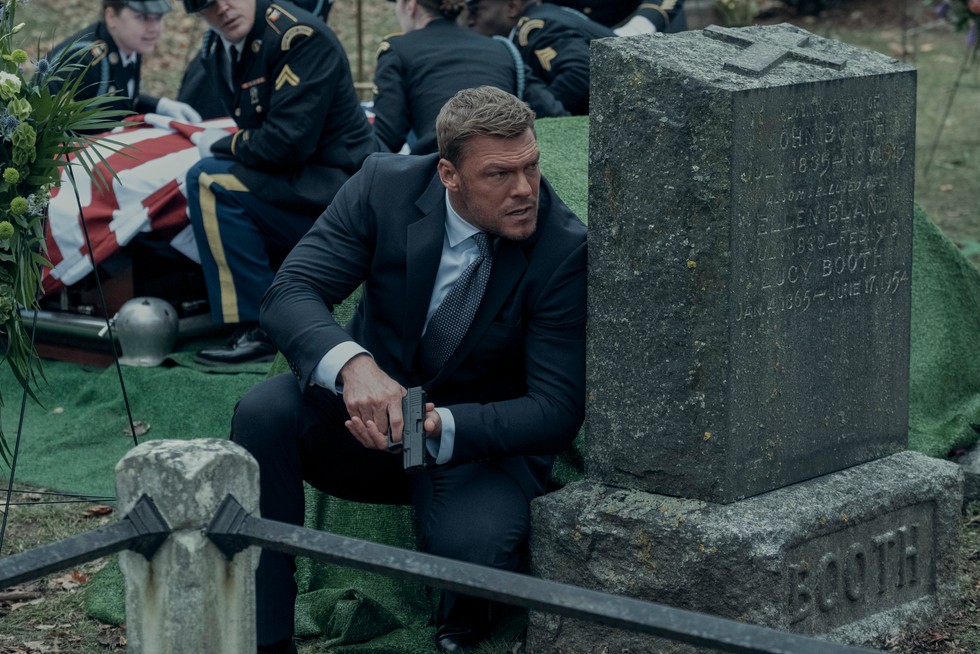 Reacher Alan Ritchson as Jack Reacher in Reacher season 2 hiding behind a tombstone, holding a pistol