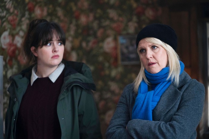 Shetland Alison O'Donnell as Tosh in Shetland, standing next to Ashley Jensen as Ruth Calder. They're stood inside a house, with Ruth looking straight ahead with her arms crossed, a stern expression on her face, and Tosh looking at her with a concerned expression on her face